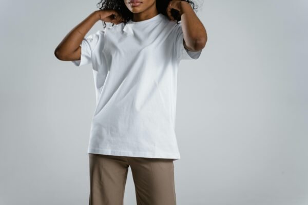 Woman wearing a white t-shirt and brown pants, adjusting her hair against a neutral background.